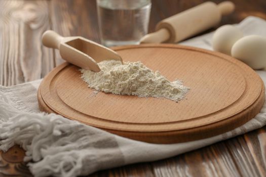 Preparations for homemade baking. Basic ingredients for baking. Flour in a wooden spoon on a cutting board. Kitchen towel, eggs, rolling pin and glass of water on a rustic wooden table