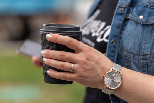 Close up of women's hands holding smartphone and a cup of coffee. A woman is dressed in a blue denim jacket, a black T-shirt and a watch on her arm