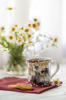 Still life with a cup of tea, cookies and daisies on a wooden background in morning sunlight