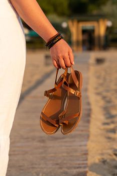 Woman holds brown leather sandals in her hand and looks at the beach hotel. Early morning at the seashore