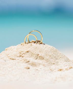 Pair of gold wedding ring bands jewelry in sand on tropical desert island beach during summer with blue ocean background