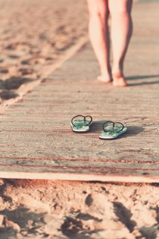 Bare feet of a girl walking on a wooden floor on the seashore at sunny summer morning. Green flip flops in the foreground.