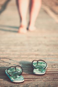 Bare feet of a girl walking on a wooden floor on the seashore at sunny summer morning. Green flip flops in the foreground.