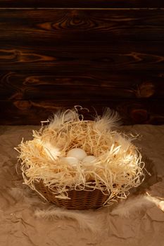 Chicken eggs lie on a wicked paper straw in a wicker basket on a dark wooden background in the morning sun