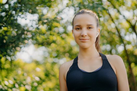 Photo of beautiful sporty woman in sportswear uses wireless earphone for listening music rests in green park looks directly at camera has rest after outdoor training involved in sport activities