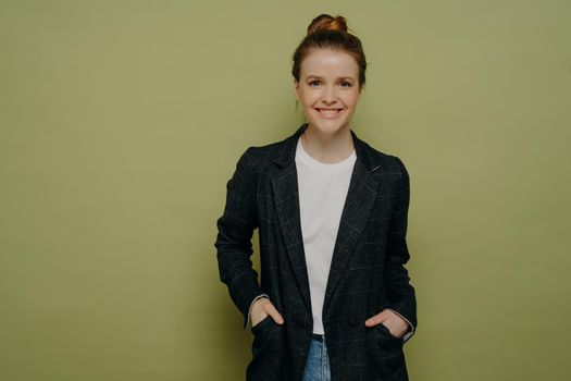 Smiling confident teenage girl with hair in bun wearing stylish plaid blazer and jeans keeping hands in pockets and looking at camera with happy expression while standing against green wall