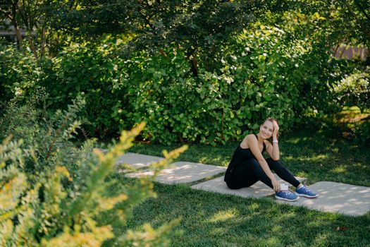 Horizontal full length shot of pleased brunette sporty woman listens motivation playlist before start running dressed in active wear poses on path uses application online. Sport and wellness concept