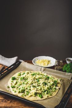 Homemade pizza with broccoli, pesto sauce, spices and cheese in a baking tray ready for baking in oven