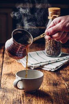 Pouring turkish coffee into white cup. Jar of roasted coffee beans on background