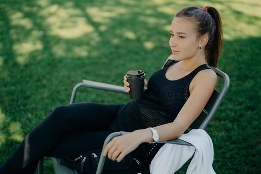Outdoor shot of thoughtful dark haired beautiful female dressed in sports clothes drinks takeaway coffee spends free time in open air poses on chair at garden enjoys good weather breathes fresh air