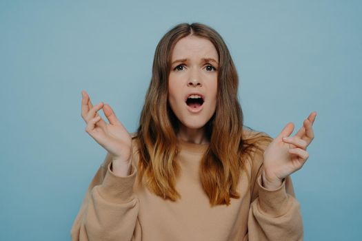 Emotional young woman with wavy ombre hair expressing shock and disbelief while looking away from camera having arms up, wearing loose casual sweater and standing against blue studio background
