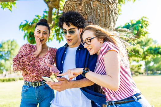 Three young friends in city park with surprised faces pointing smartphone enjoying social networks on mobile cell. Young people having fun together in nature with wi-fi internet connection technology