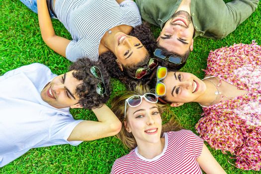 Shot from above of five happy multiracial friends lying head to head with colored sunglasses looking at camera from bottom. Mixed race group of smiling cute young people having fun together in nature
