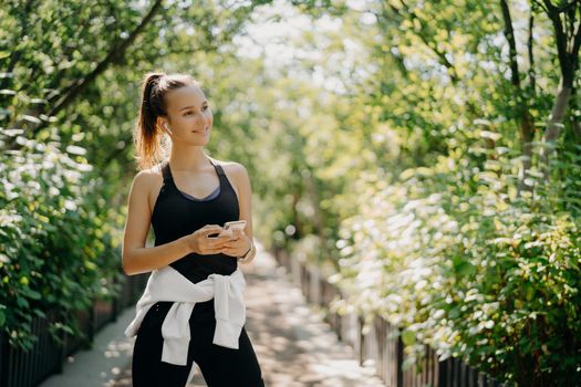Horizontal shot of active sporty woman uses smartphone for checking results after jogging dressed in active wear enjoys warm sunny day listens music via earphones. Healthy lifestyle concept.