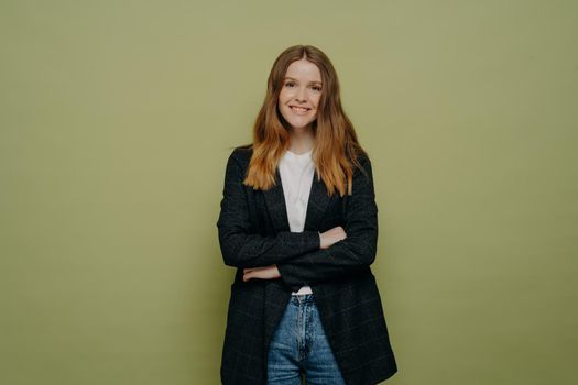 Happy young woman dressed in stylish casual outfit with crossed arms looking at camera and smiling gladfully while posing in studio, pleasant female student standing against green background