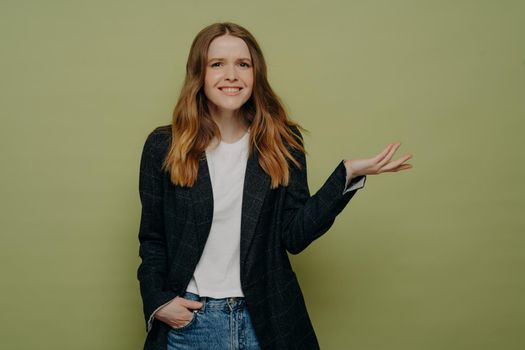 Smiling young woman with tipping hand demonstrating confusion and uncertainty wearing dark formal jacket, white top and jeans posing isolated over grey studio wall with one hand in pocket