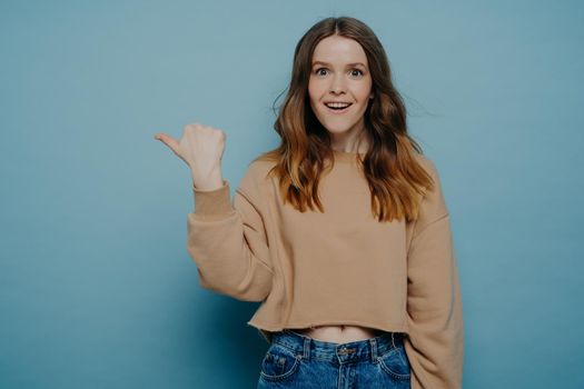 Amused pretty young female with ombre hair in loose casual light brown sweater and jeans pointing her finger at something attracting attention, standing against blue wall. Advertisement and promotion