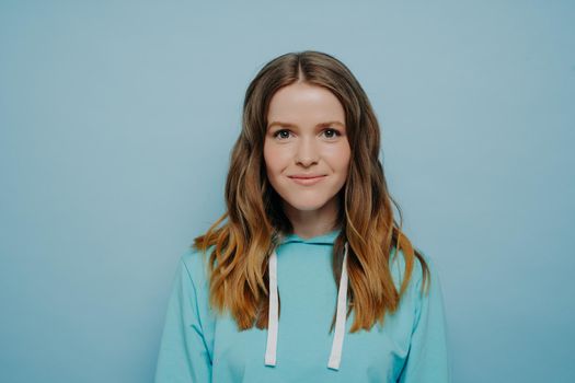 Portrait of teenager girl with wavy ombre hairstyle smiling while looking at camera in comfortable casual blue sweatshirt standing against studio background