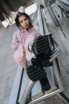 Young woman with futuristic looks. Girl with black and white dreadlocks or pigtails. Against the background of a futuristic building. Focus on shoes