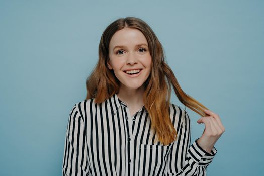 Positive smiling young girl in stripy black and white blouse demonstrating curiosity and amusement while holding strand of wavy brown hair posing over light blue background