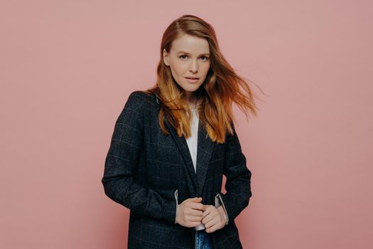 Medium shot of attractive woman with ginger hair looking at camera wearing dark formal jacket and white top posing isolated over pink studio background. Young people concept