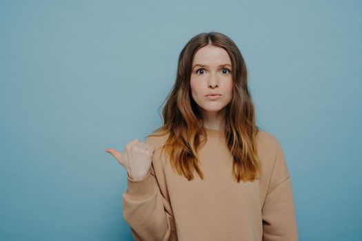 Surprised teenage girl in casual outfit pointing aside with finger at copy space and making shoked face expression while advertising product or saying about unbelievable sale,, isolated over blue wall