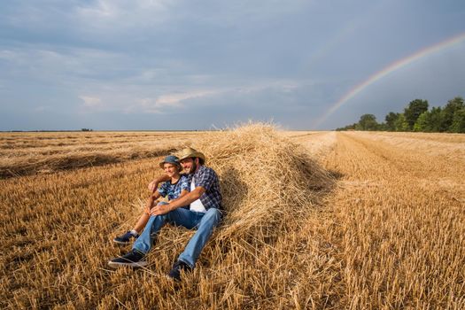 Father and son are resting after successful harvest. Rainbow in the sky behind them.