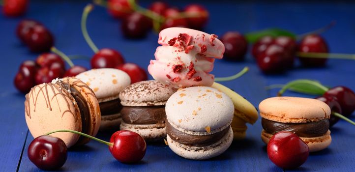 multicolored macarons and ripe red cherries on blue wooden background, close up