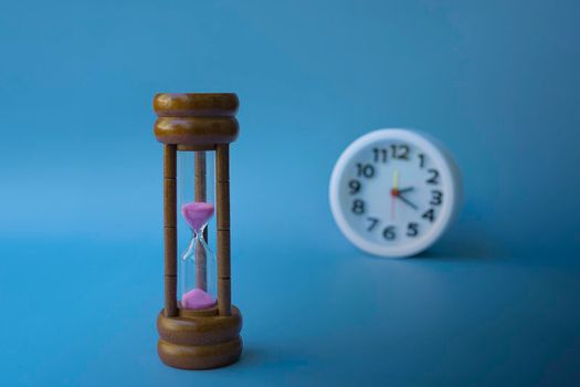 Hourglass as time passing concept for business deadline and running out of time with blue background and blurred alarm clock. Space for text