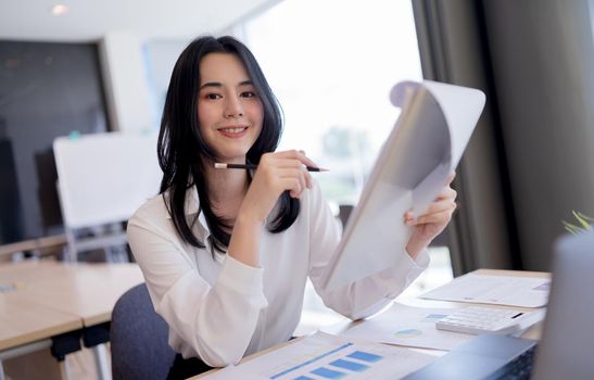Beautiful Asian business lady holding paperwork, pencil and smiling looking at camera while working in bright modern office.