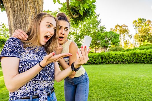 Two girls best friends look amazed at smartphone with disbelief grimaces and wide-open mouths and eyes in surprise Young women couple having fun with technology enjoying social network in a city park