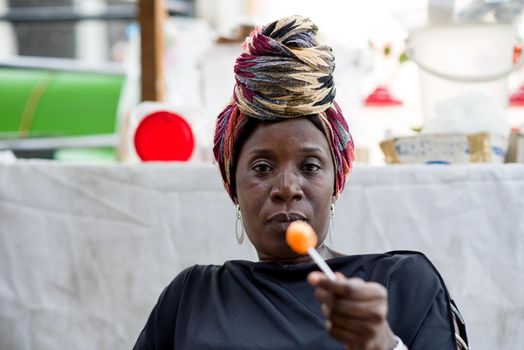 Woman with head scarf sitting behind her sales stand is holding a licked lollipop in her hand.