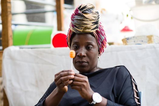 Woman with head scarf sitting behind her sales stand is holding a licked lollipop in her hand.