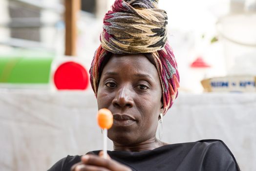 Woman with head scarf sitting behind her sales stand is holding a licked lollipop in her hand.