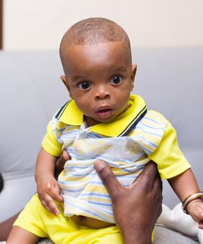 Close up of baby in his dad's arms sitting in the couch at home watching the camera surprised