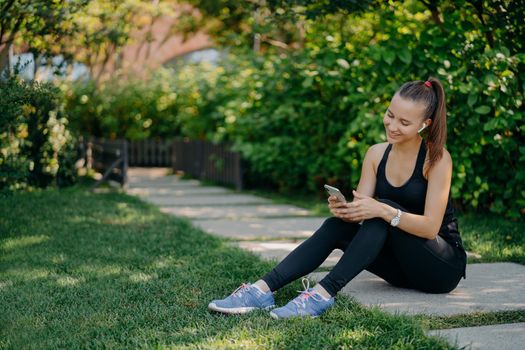 Pleased athletic woman in sportsclothes smiles cheerfully surfs mobile phone while listens music via wireless earphones takes break after sunny morning workout uses app for activity tracking