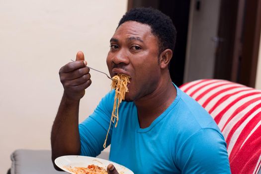 young man sitting in an armchair eating spaghetti holding a plate in his hand.