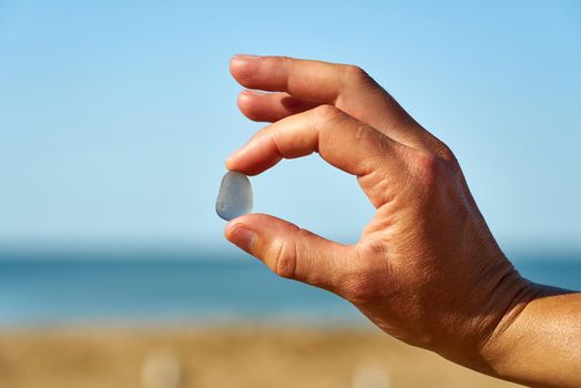 A blue piece of sea glass is held up with the sand, sea and sky in the background.