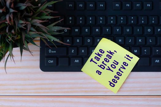 Text on yellow note with keyboard and potted plant on wooden table. Take a break, you deserve it