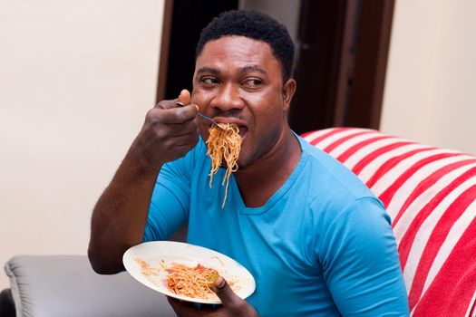 young man sitting in an armchair eating spaghetti holding a plate in his hand.